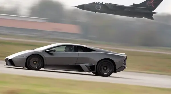 Szary Lamborghini Reventon pędzi po pasie startowym, gdy myśliwiec Tornado przelatuje nad nim.