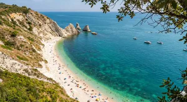 Spiaggia affollata ai piedi del Monte Conero, con acque cristalline e barche all'orizzonte.