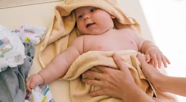 Söt bebis i handduk, kanske med tyst reflux bebis symtom. Bredvid ligger kläder och blöjor.