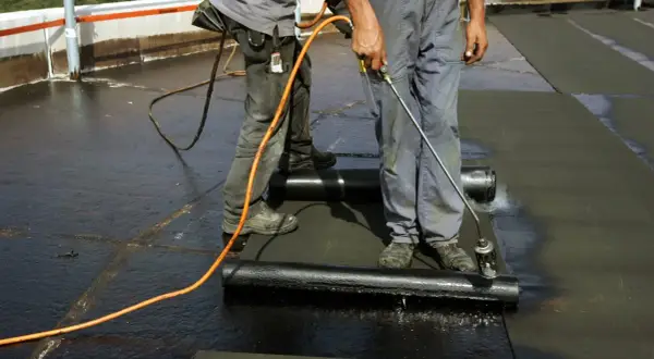 Zwei Dachdecker arbeiten auf einem Flachdach. Einer hält einen Brenner, um die Dachpappe zu schweißen.