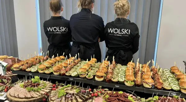 Trzy kobiety w czarnych fartuchach z logo "WOLSKI CATERING" stoją za stołem pełnym przekąsek. To idealna impreza z pysznym jedzeniem.