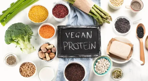 Tablica z napisem "VEGAN PROTEIN" otoczona miseczkami z roślinnymi źródłami białka: soczewicą, fasolą, orzechami, tofu, brokułem i szparagami.