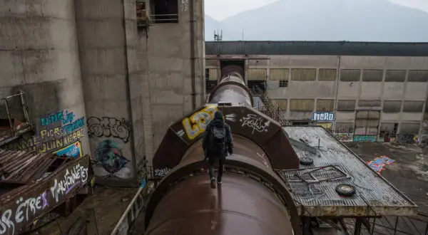 Explorateur en urbex, marchant sur un immense tuyau rouillé, traversant un grand vide industriel. Graffiti sur les murs.