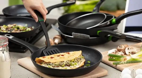 Eine Hand wendet mit einem Pfannenwender einen gefüllten Pfannkuchen in einer beschichteten Pfanne. Mehrere Pfannen und Zutaten wie Pilze und Eier sind im Hintergrund zu sehen.