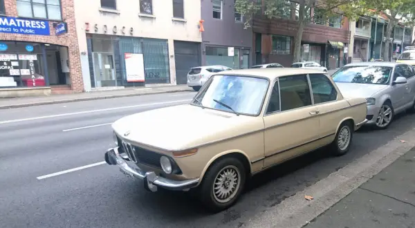 Klasyczny beżowy BMW E10 z lat 70. stoi zaparkowany przy ulicy, obok nowszego srebrnego samochodu.