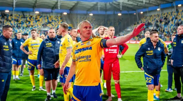 Piłkarze w żółto-niebieskich strojach, symbolizujący polskie kluby piłkarskie, stoją na boisku. Jeden z nich gestykuluje, przemawiając do drużyny.