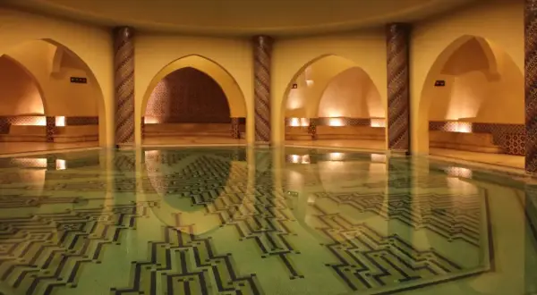 Wnętrze hammamu z basenem o geometrycznym wzorze na dnie. Wokół łuki, kolumny zdobione mozaiką i kamienne ławy.