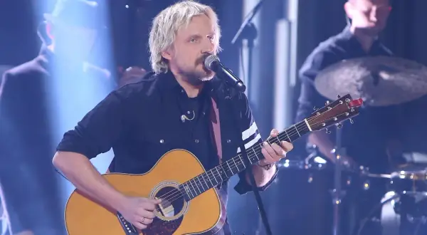 Artysta z blond włosami gra na gitarze akustycznej, śpiewając do mikrofonu. Juz teraz wiem ze dni tekst.