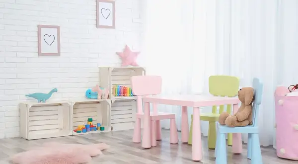 Kolorowy stolik z krzesełkami dla dzieci, idealny do zabawy i nauki. Na ścianie wiszą ramki z serduszkami, a obok stoją drewniane skrzynki z zabawkami.