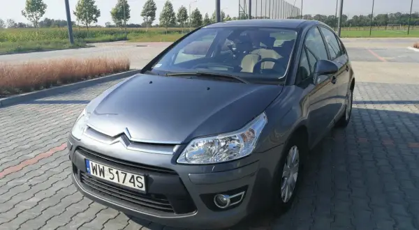 Szary Citroen C4 z 2008 roku na parkingu z kostki brukowej, w tle drzewa i boisko.