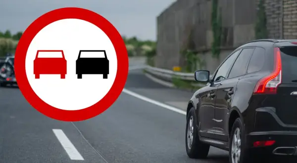 Znak zakazu wyprzedzania na podwójnej ciągłej. Czarny SUV jedzie po drodze, a na pierwszym planie znak drogowy.