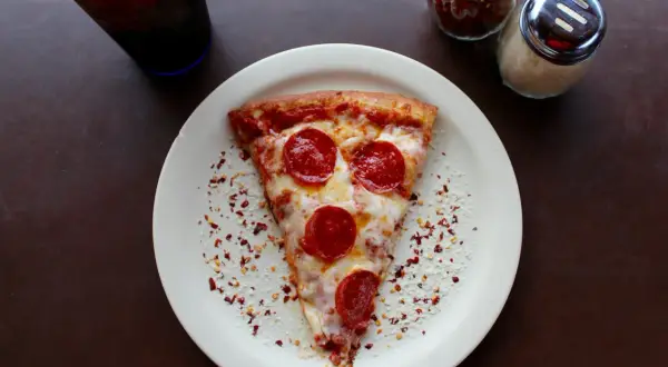 Kawałek pizzy pepperoni na talerzu, obok słoiczki z przyprawami i napój. Zastanawiasz się, ile kosztuje pizza we Włoszech?