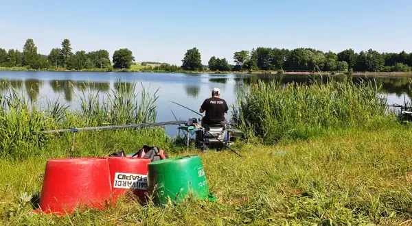 Wędkarz z napisem "FEBI FISH" na koszulce siedzi nad spokojnym stawem, gotowy do łowienia.