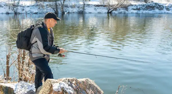 Mann angelt bei Minusgraden am Flussufer, das mit Schnee bedeckt ist. Er trägt eine Jacke und einen Rucksack.