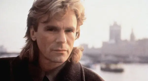 MacGyver obsada: Richard Dean Anderson jako MacGyver, z charakterystyczną fryzurą i w płaszczu, na tle mglistego miasta.