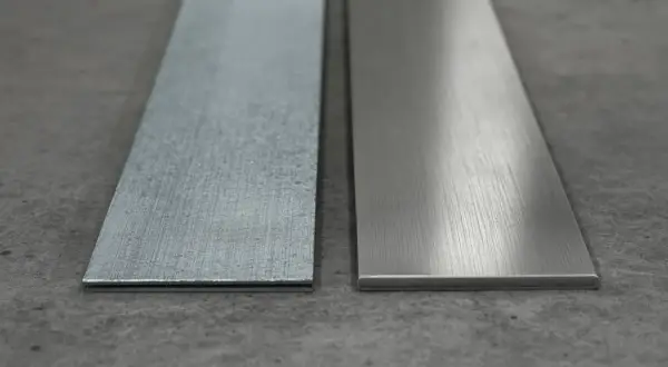 Dwa metalowe paski leżą obok siebie na betonowym podłożu. Jeden jest szorstki, drugi gładki, jakby przygotowany do montażu kotwy.