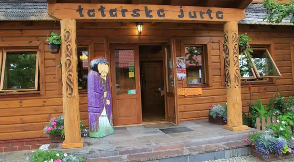 Drewniana fasada z napisem "tatarska jurta". Przed wejściem stoi kolorowa rzeźba postaci w stroju tatarskim.