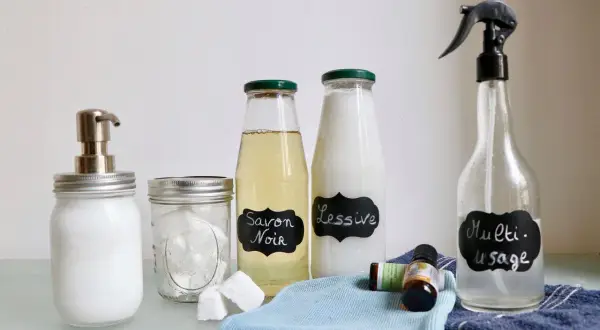 Bouteilles étiquetées "Savon Noir", "Lessive" et "Multi-usage", ainsi qu'un distributeur de savon, des pastilles et des huiles essentielles. Des produits faits maison pour un nettoyage écologique.