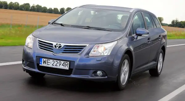 Szary Toyota Avensis T27 1.8 benzyna w kombi jedzie po drodze, za nim pola.