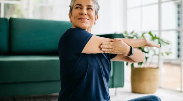 Une femme souriante s'étire, prouvant qu'il est possible de redresser son dos à 60 ans. Elle est assise sur un tapis, près d'un canapé vert.