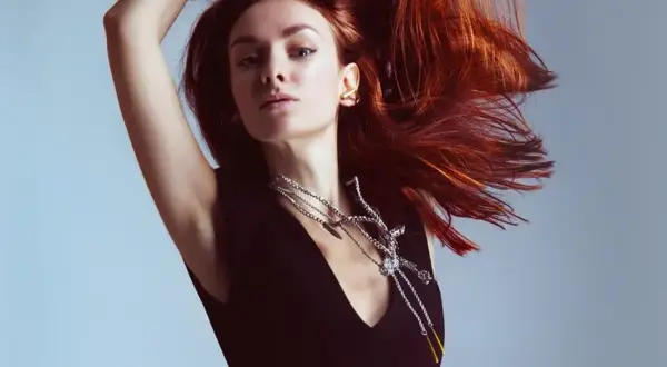 Mujer con cabello cobrizo vibrante, moviéndose con gracia. Luce un collar plateado llamativo y un pendiente dorado.