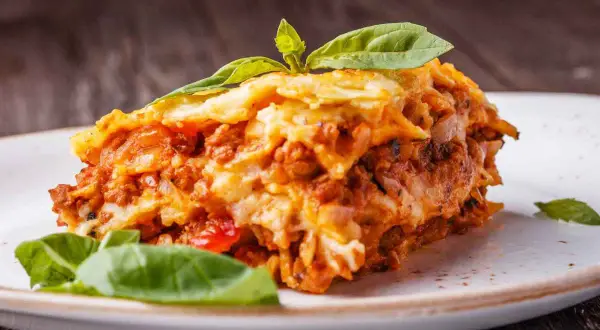Una fetta di lasagna salentina, ricca e saporita, guarnita con basilico fresco, su un piatto bianco.
