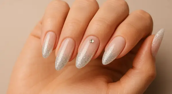 Main d'une femme aux ongles en amande, ornés d'un dégradé de paillettes argentées et d'un petit strass sur l'annulaire.