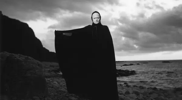En skrämmande gestalt med vit ansiktsmålning och svart kåpa står på en stenig strand. En ikonisk scen från filmer med Ingmar Bergman.