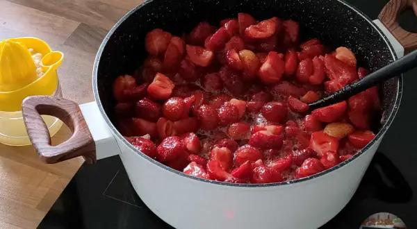 Duży garnek z gotującymi się truskawkami na kuchence indukcyjnej. Obok żółta wyciskarka do cytrusów.