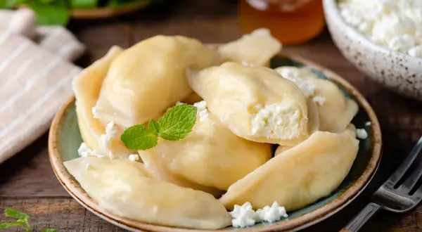 Pyszne pierogi z serem, udekorowane miętą i posypane serem. Obok słoiczek miodu i miseczka z serem.
