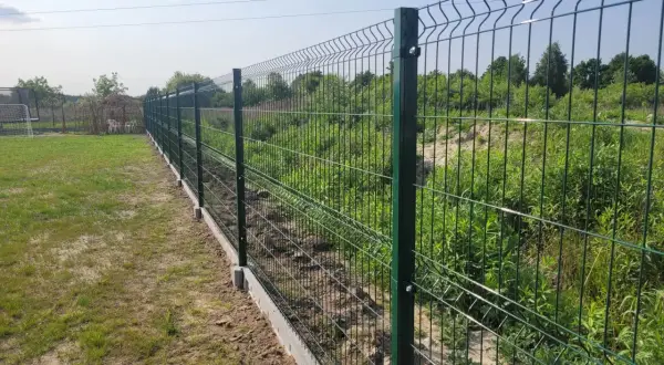 Nowe, zielone ogrodzenie działki rekreacyjnej z betonowym podmurowaniem. W tle trawa i zarośla.