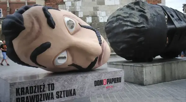 Dwie gigantyczne głowy, jedna jasna, druga ciemna, sugerują, z jakiego materiału można wykonać rzeźbę. Napis "Dom z papieru" informuje o pochodzeniu.