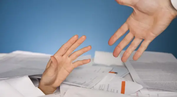 Dłoń wyciągnięta z papierów, druga dłoń oferuje pomoc. Symbolizuje trudności i potrzebę wsparcia w procesie restrukturyzacji.