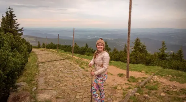 Uśmiechnięta turystka z kijkami na kamienistej ścieżce. Widok na góry i doliny. Czas wejścia na Śnieżkę zależy od kondycji i wybranej trasy.