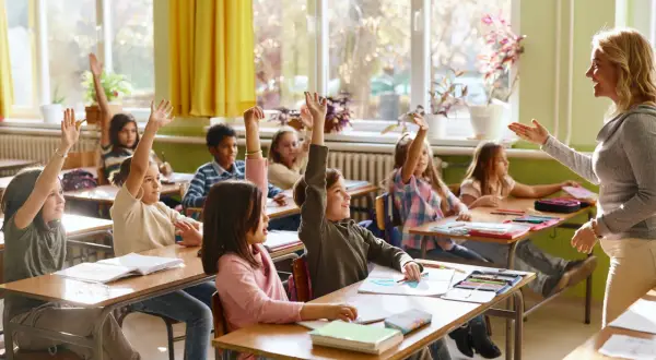 Grundschüler mit erhobenen Händen, bereit zu antworten. Die Sitzordnung zur Wand hin ist gut erkennbar, die Lehrerin lächelt.