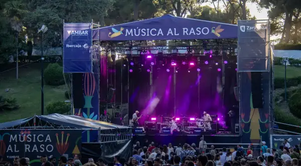 Escenario iluminado con luces púrpuras para un concierto de **música al raso**. Una multitud disfruta del espectáculo al aire libre.