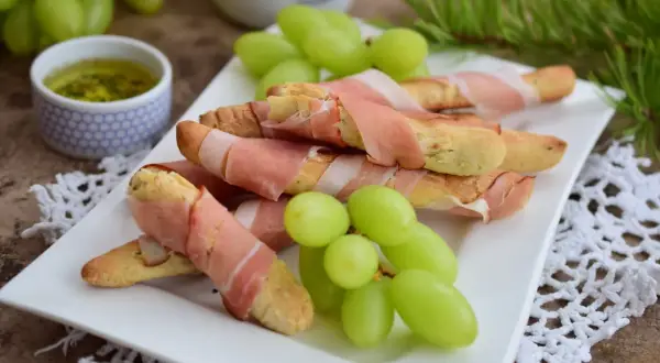 Grissini z szynką parmeńską, zielone winogrona i sos na talerzu. Idealna przekąska na przyjęcie.
