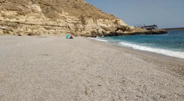 Arena y guijarros en la playa de las salinicas, con acantilados dorados y mar azul. Una sombrilla azul protege a unos bañistas.