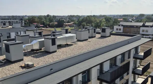 Widok na płaski dach z systemem wentylacyjnym i żwirem. Widoczne są warstwy stropodachu, elementy wentylacyjne i fragmenty budynków mieszkalnych.