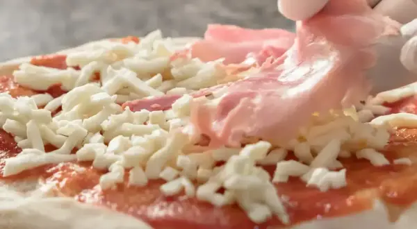 Rękawiczka w białej rękawiczce dodaje cienkie plastry różowej szynki na pizzę, która jest już pokryta sosem pomidorowym i startym serem.