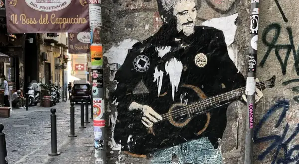 Graffiti di Pino Daniele con la chitarra, un omaggio a Napoli. I testi "Resta... resta cu'mmè" risuonano tra i vicoli.