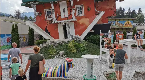 Ein rotes Haus steht auf dem Kopf, die Treppe führt ins Nichts. Eine Familie bestaunt das Spektakel, bunte Skulpturen und ein Dino-Land im Hintergrund.