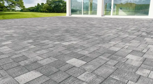 Un ampio patio in gres carrabile grigio, con pavimentazione a motivi geometrici, si estende davanti a una moderna casa con vetrate.