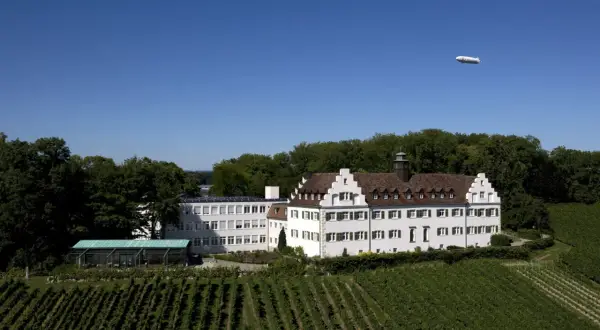 Schloss Hersberg, umgeben von Weinbergen, mit einem Zeppelin am blauen Himmel.