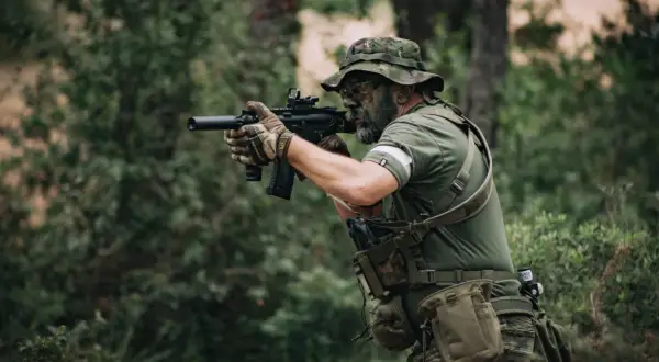 Jugador de deporte airsoft con camuflaje facial y equipo táctico, apuntando con su réplica de rifle en un bosque.
