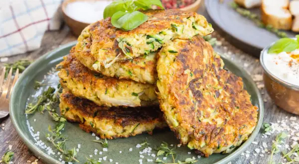 Goldbraune Zucchinipuffer mit Haferflocken, garniert mit Basilikum, auf einem Teller mit Kräutern und grobem Salz.