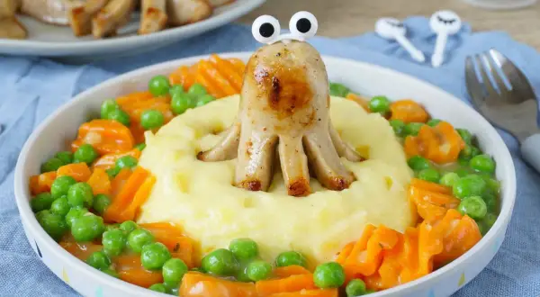 Ein lustiger Tintenfisch aus Bratwurst vom Vortag, der aus Kartoffelbrei mit Erbsen und Karotten schaut.
