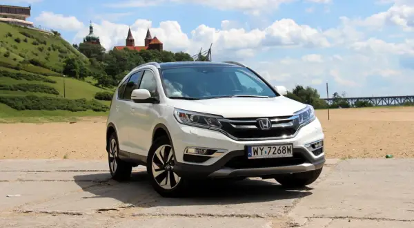 Biała Honda CR-V 2.0 benzyna na tle piaszczystego brzegu i zielonego wzgórza z budynkami.