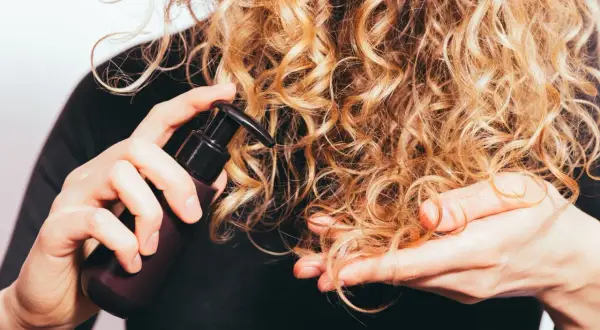Delikatna pielęgnacja włosów wysokoporowatych. Ręka z buteleczką kosmetyku aplikuje go na blond loki.