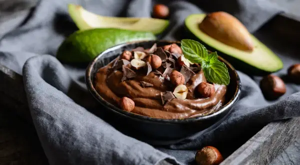 Kremowa nutella z awokado, udekorowana orzechami laskowymi, kawałkami czekolady i miętą. Obok leżą połówki awokado.